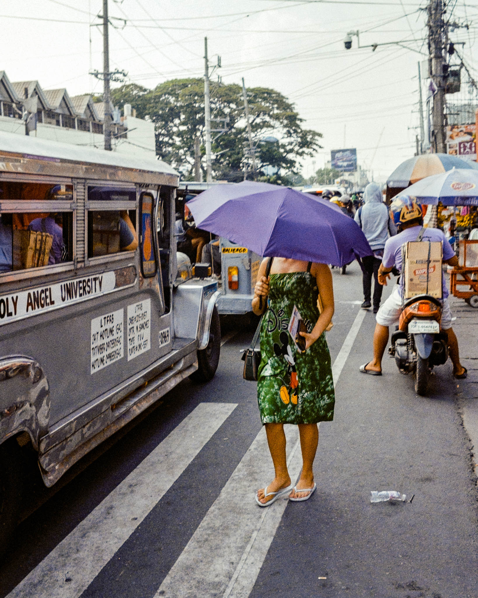 Woman with purple umbrella waiting amid bustling traffic, including a jeepney and tricycle, in the Philippines.