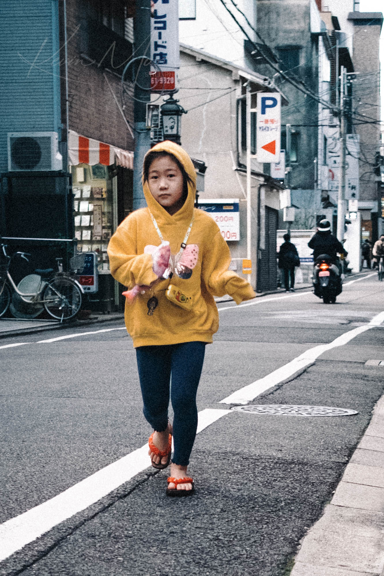 Youth in yellow hoodie strolling on busy Japanese city street with pink items in bag.