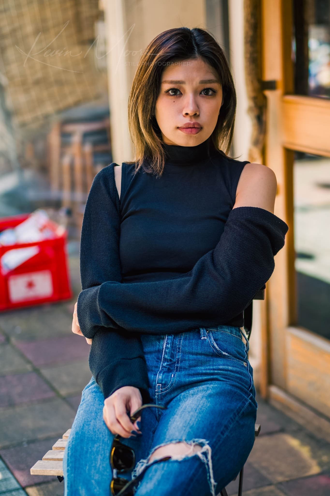 Young woman in black turtleneck and ripped jeans sitting on a stool in urban setting.
